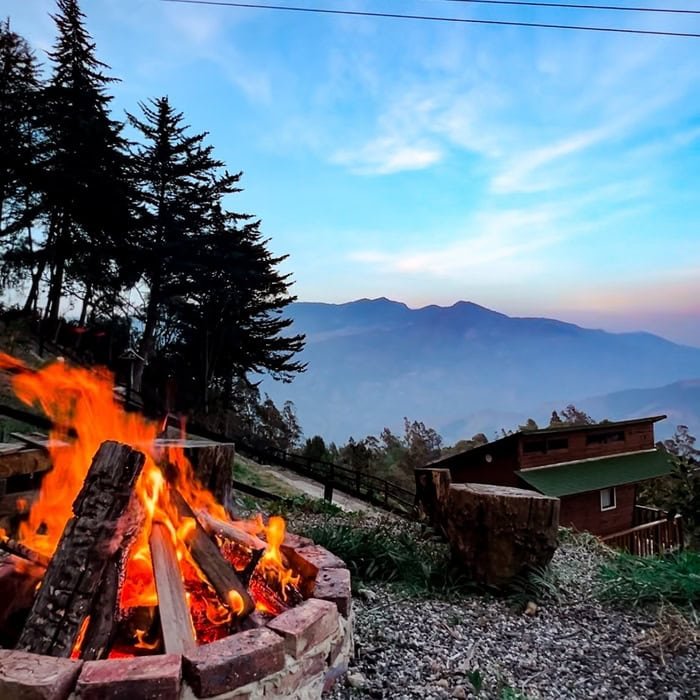 Fogata encendida con árboles y montañas de fondo, cielo azul con nubes